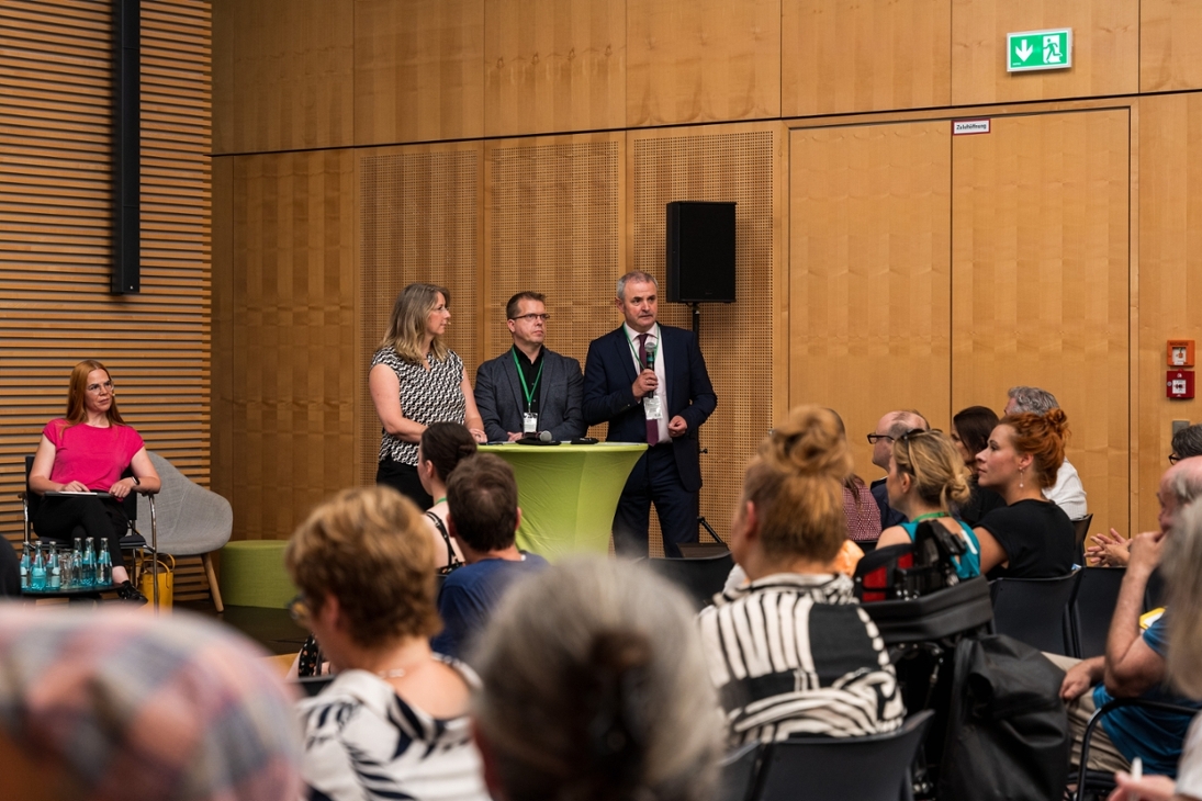 Der Vorsitzende des Landesbeirats für Inklusion der Menschen mit Behinderungen, Ralph Beckert und der Landesinklusionsbeauftragte Michael Welsch stehen zusammen mit der Moderatorin Melanie Sterczewski vor dem Publikum des Wahlforums an einem Stehtisch.