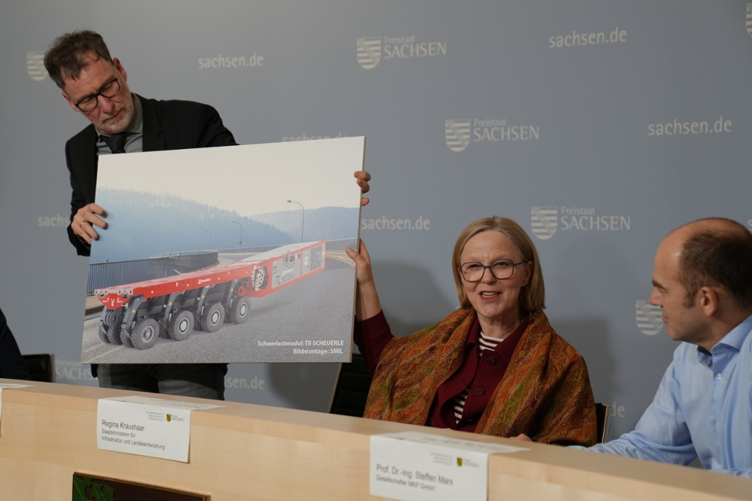 Abteilungsleiter für Mobilität, Stephan Berger, hält Fotomontage hoch. Darauf zu sehen ist die gesperrte Brücke in Bad Schandau und darauf, die dort bald fahrenden ferngesteuerten Schwerlastmodule. Staatsministerin Regina Kraushaar erklärt das Verfahren.