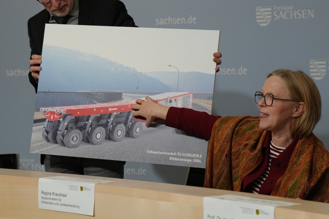 Abteilungsleiter für Mobilität, Stephan Berger, hält Fotomontage hoch. Darauf zu sehen ist die gesperrte Brücke in Bad Schandau und darauf, die dort bald fahrenden ferngesteuerten Schwerlastmodule. Staatsministerin Regina Kraushaar erläutert die Vorgehensweise.