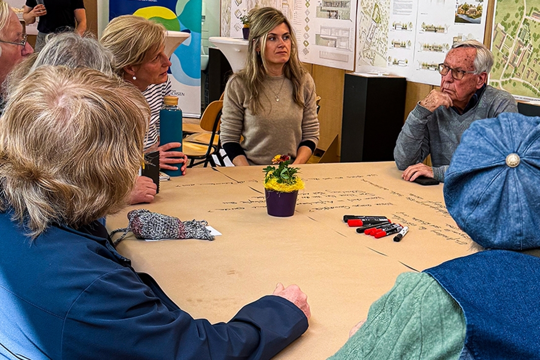 Ein Gruppe von Menschen sitzt in einer Diskussionsrunde an einem großen Tisch in kreativer Arbeitsatmosphäre zusammen. Auf dem Tisch liegen Filzstifte und Pergament-Papier auf dem Ideen und Gedanken der Teilnehmer notiert sind. Im Hintergrund sind Architektur-Entwurfe an Pinnwänden zu sehen.