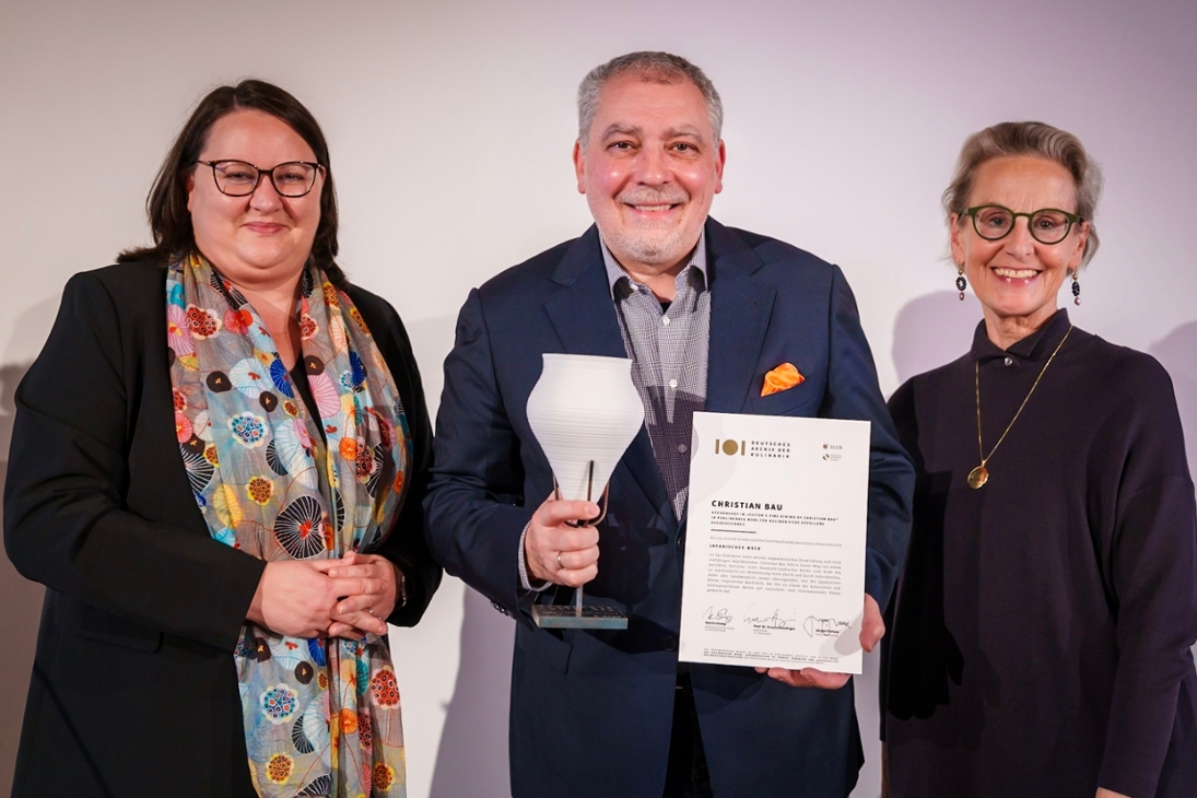 Katrin Stump (links) und Prof.in Ursula Staudinger (rechts) übergeben die Urkunde und Gefäßstatuette an Spitzenkoch Christian Bau.
